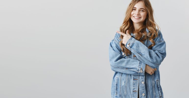 Girl points at her director, telling rumor about him. Positive charming caucasian female office worker in denim jacket pointing at upper left corner or behind while smiling broadly, being pleased